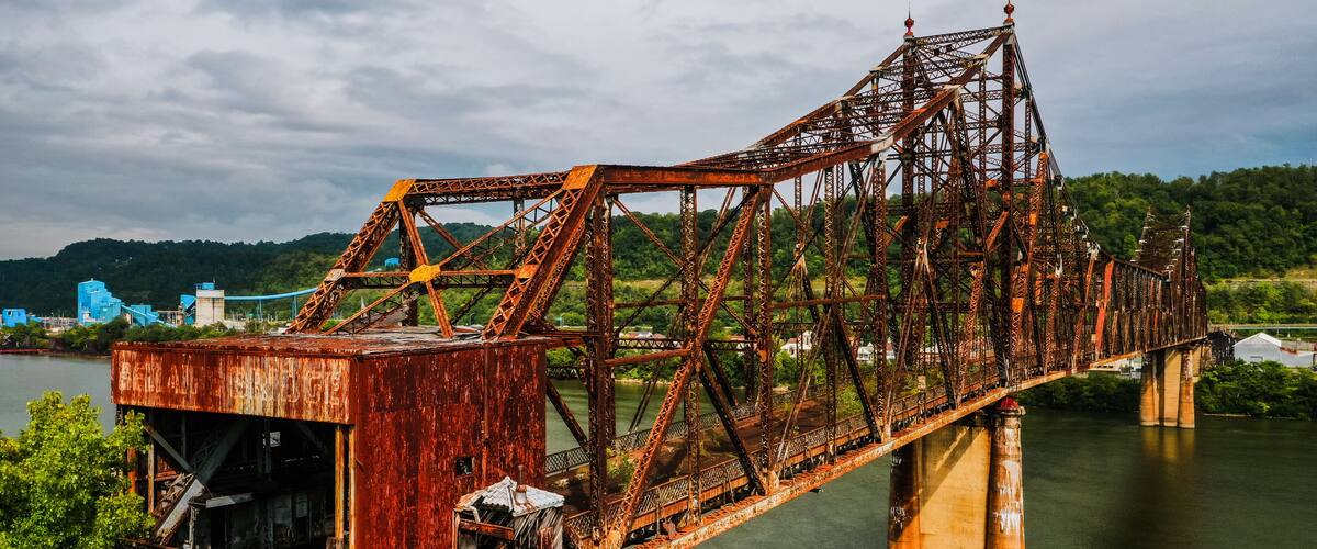 A rusted steel bridge over a river with a forested hillside in the background.