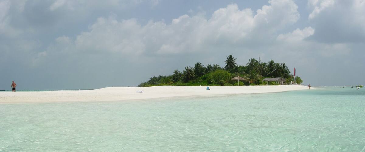 A beach with clear turquoise water, a white sandy shore, and a small island with palm trees and a hut.