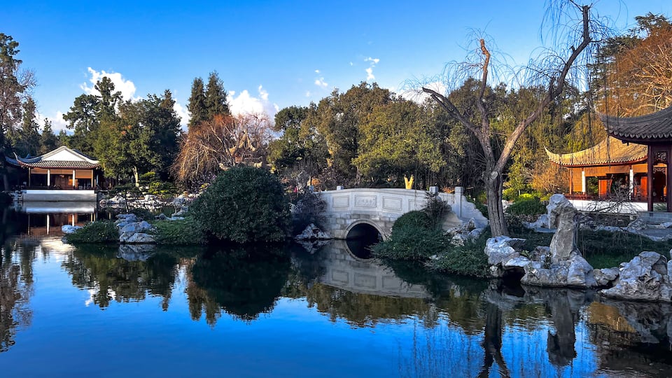 A serene pond with a stone bridge, traditional architecture, and lush trees.