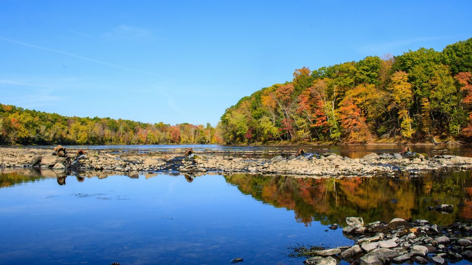 A calm river with clear reflections, surrounded by autumn-colored trees and rocky shores.