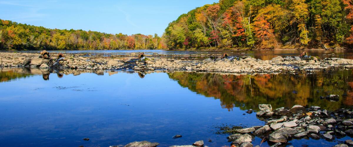 A calm river with clear reflections, surrounded by autumn-colored trees and rocky shores.