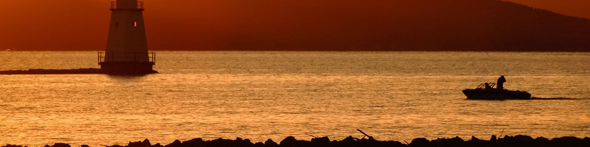 Golden sunset on Lake Champlain looking west towards the Adirondack Mountains of New York. Lighthouse and boat with fishermen in silhouette.