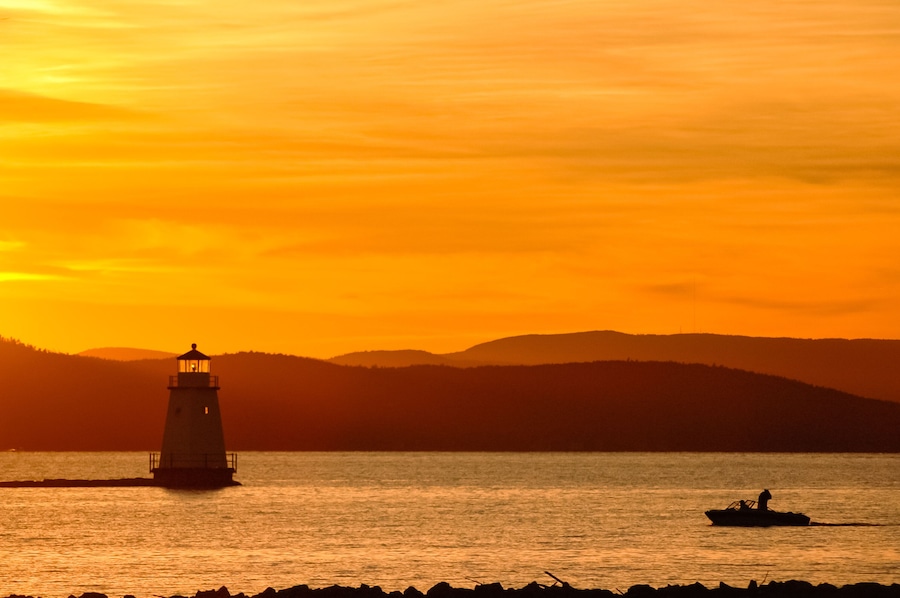 Golden sunset on Lake Champlain looking west towards the Adirondack Mountains of New York. Lighthouse and boat with fishermen in silhouette.