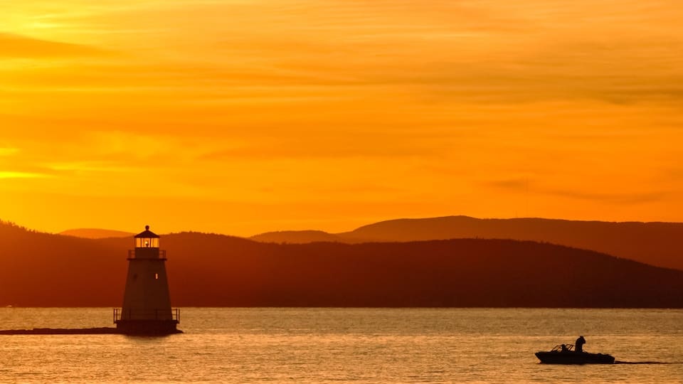 A lighthouse at sunset, a boat on the water, and a hilly landscape in the background.