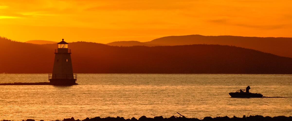 A lighthouse at sunset, a boat on the water, and a hilly landscape in the background.