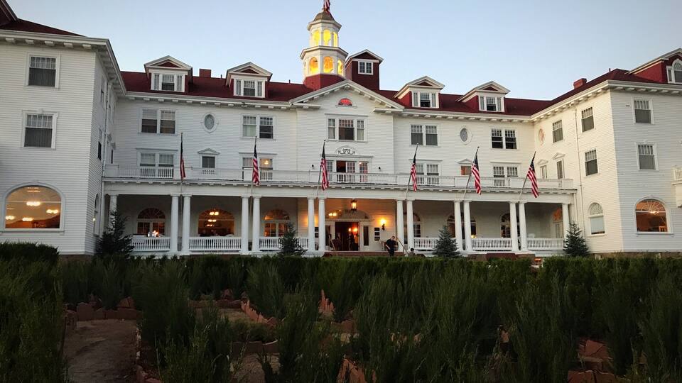 A large white building with multiple balconies and flags, a tower with a flag, and a well-maintained garden in front.