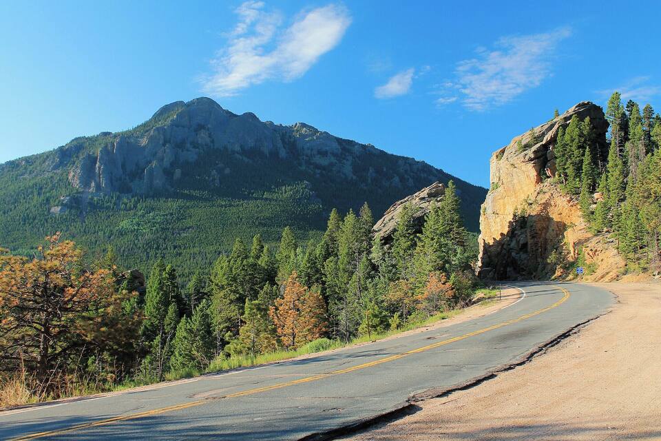 A winding mountain road with a rocky outcrop on the right and a forested mountain in the background.