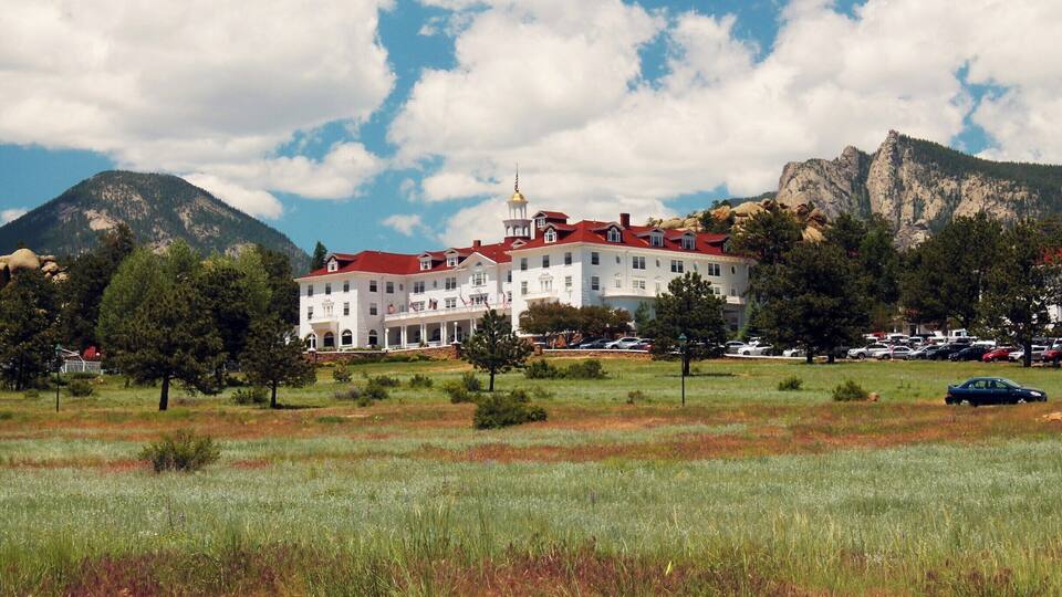 A large white building with a red roof, surrounded by a grassy field and trees, with mountains in the background.