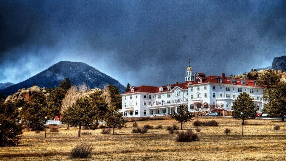 A large white building with a red roof, surrounded by trees and mountains in the background.