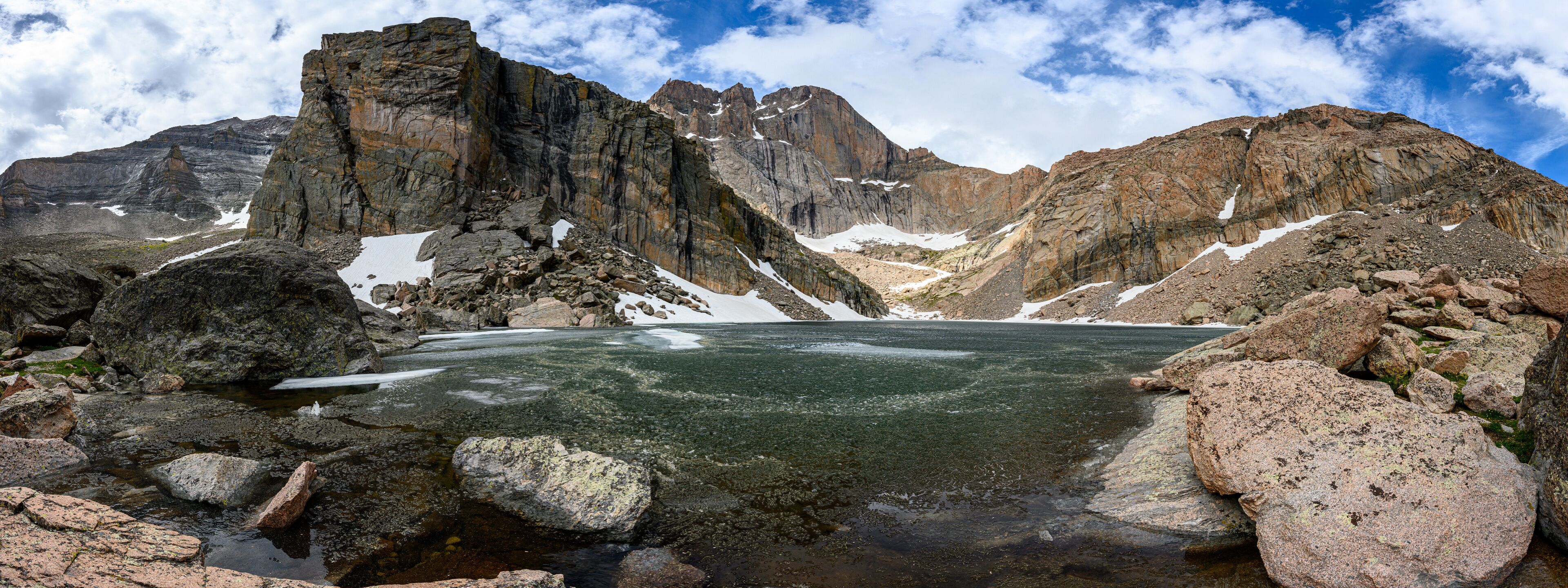 A mountainous landscape with a glacial lake surrounded by rocky terrain and snow patches.