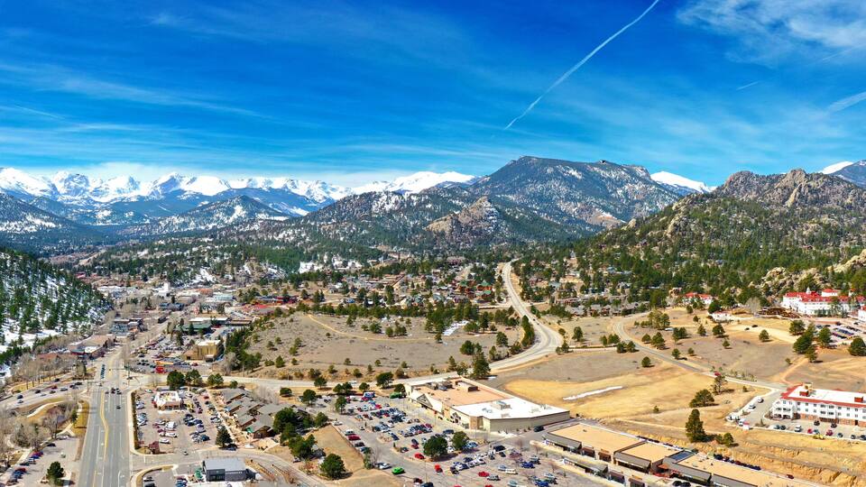 A town nestled between snow-capped mountains, with a mix of residential and commercial buildings, roads, and a clear blue sky.