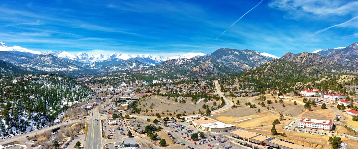 A town nestled between snow-capped mountains, with a mix of residential and commercial buildings, roads, and a clear blue sky.