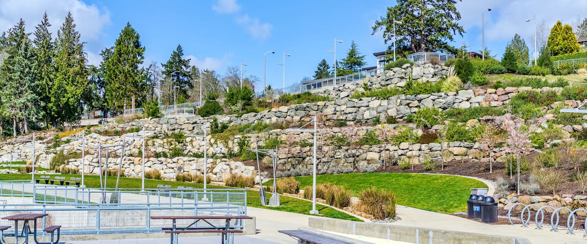 A park with picnic tables, a stone retaining wall, and a grassy hill with trees.