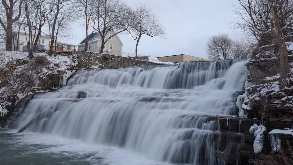 A waterfall cascading over rocky terrain with snow-covered trees and houses in the background.