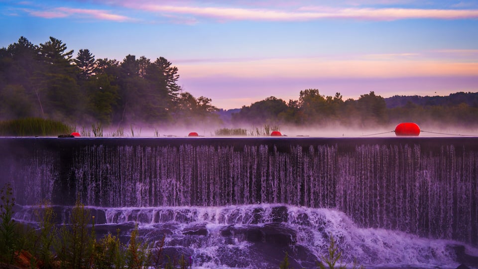 A waterfall with red buoys, surrounded by trees and a misty sky.