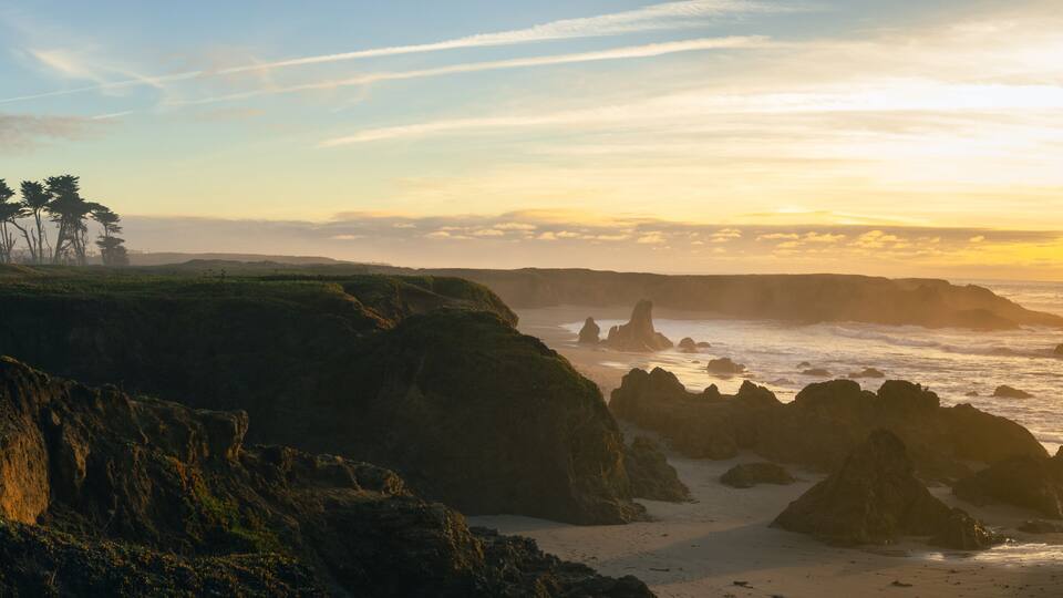 A coastal landscape with rocky cliffs, a sandy beach, and a group of trees.
