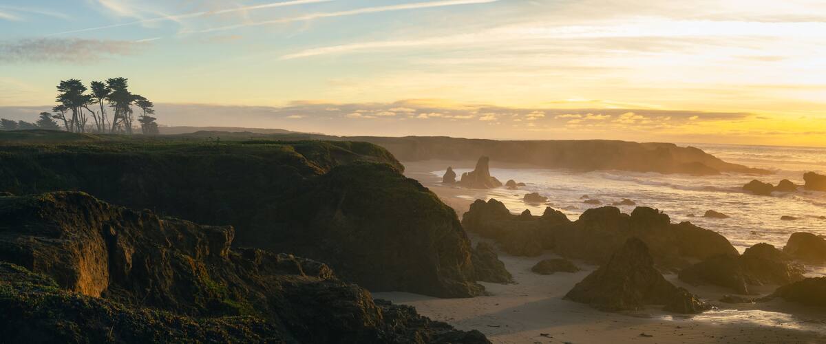 A coastal landscape with rocky cliffs, a sandy beach, and a group of trees.