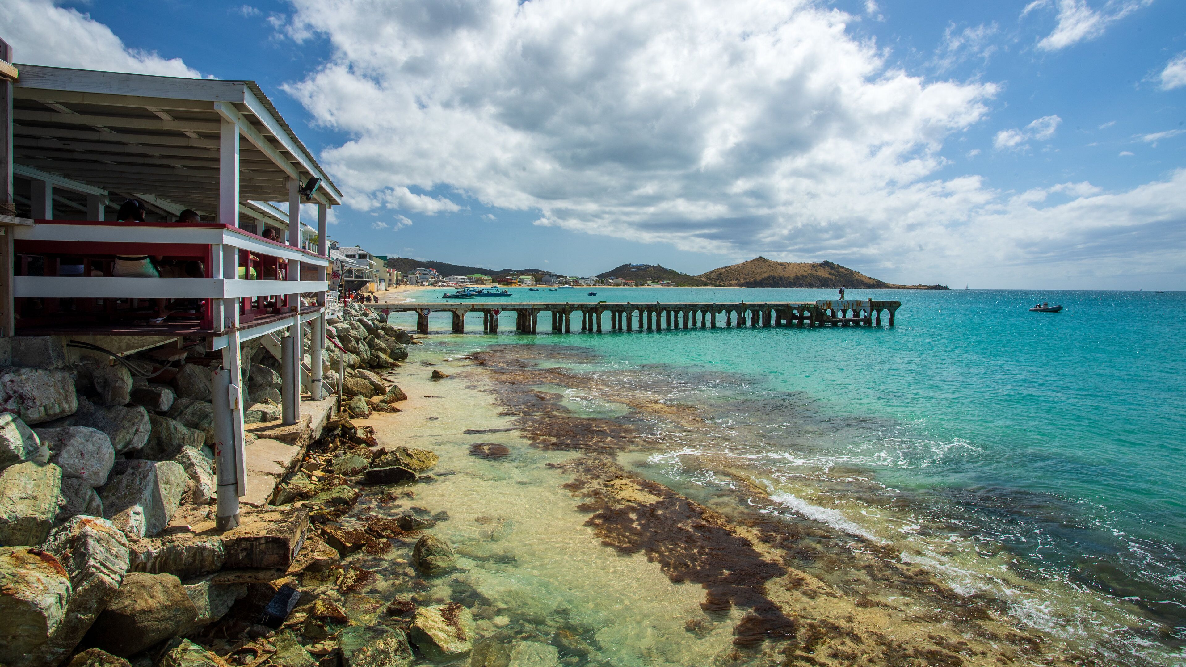 A coastal scene with a pier extending into turquoise waters, rocky shoreline, and a distant hill.