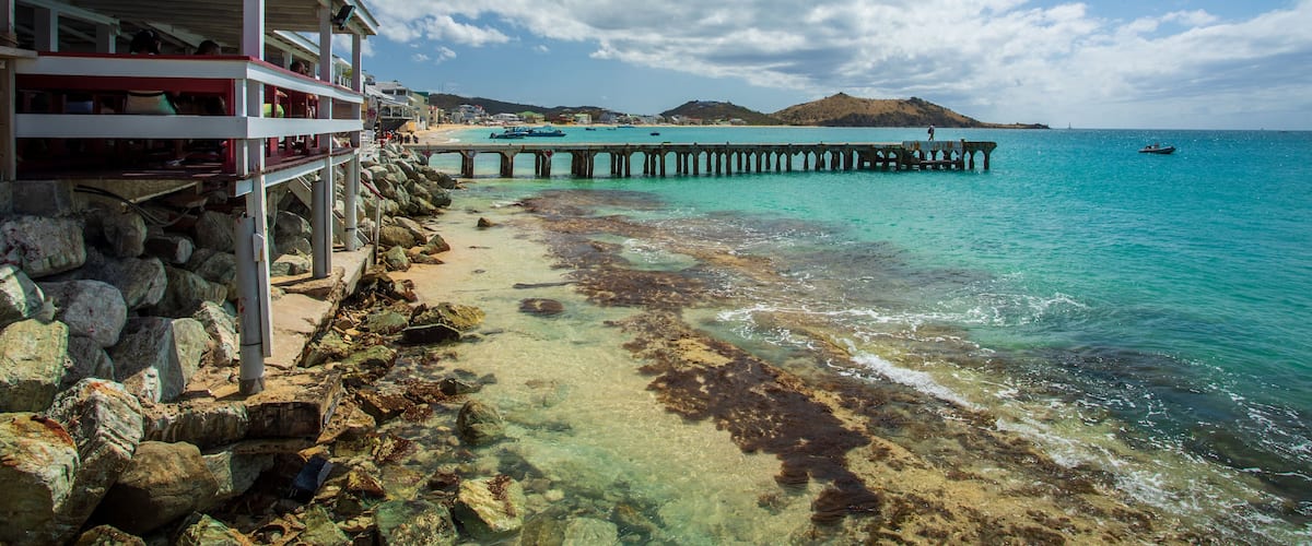 A coastal scene with a pier extending into turquoise waters, rocky shoreline, and a distant hill.