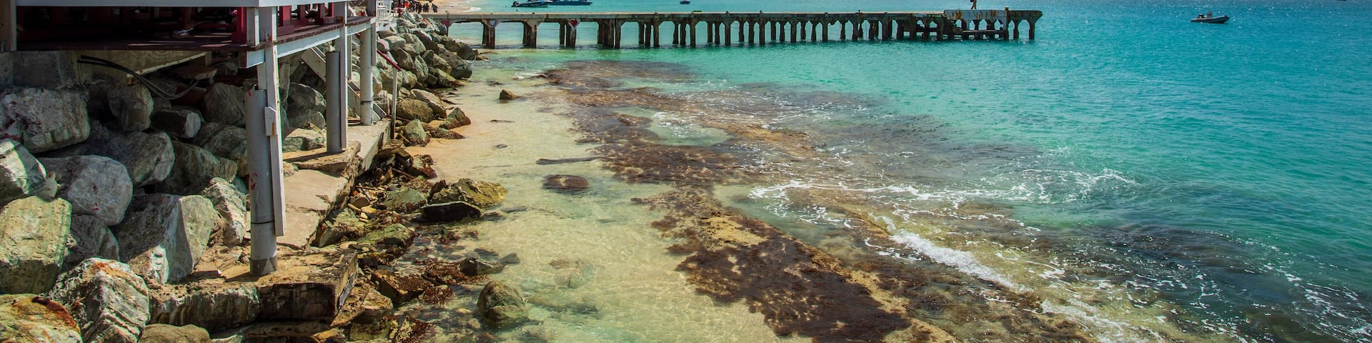 A coastal scene with a pier extending into turquoise waters, rocky shoreline, and a distant hill.