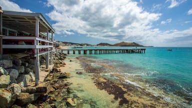 Escena costera con un muelle que se adentra en aguas turquesas, costa rocosa y una colina distante.