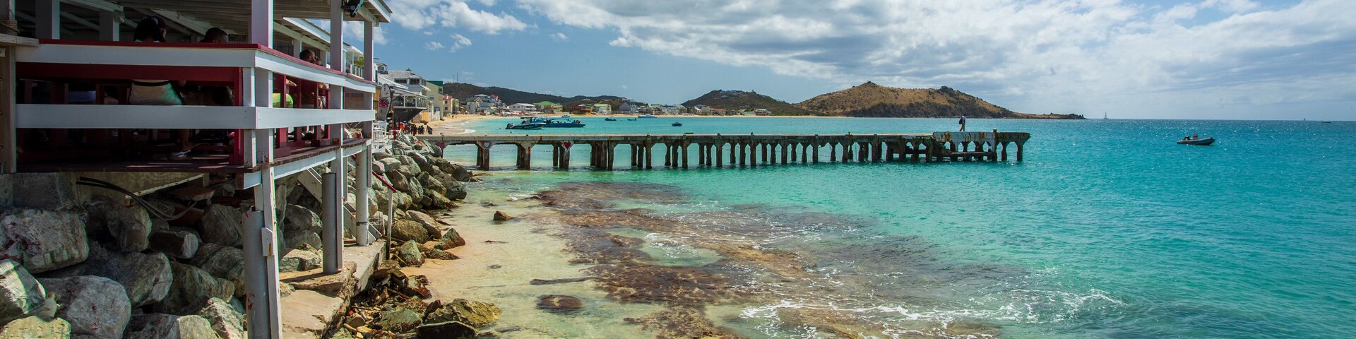 Escena costera con un muelle que se adentra en aguas turquesas, costa rocosa y una colina distante.