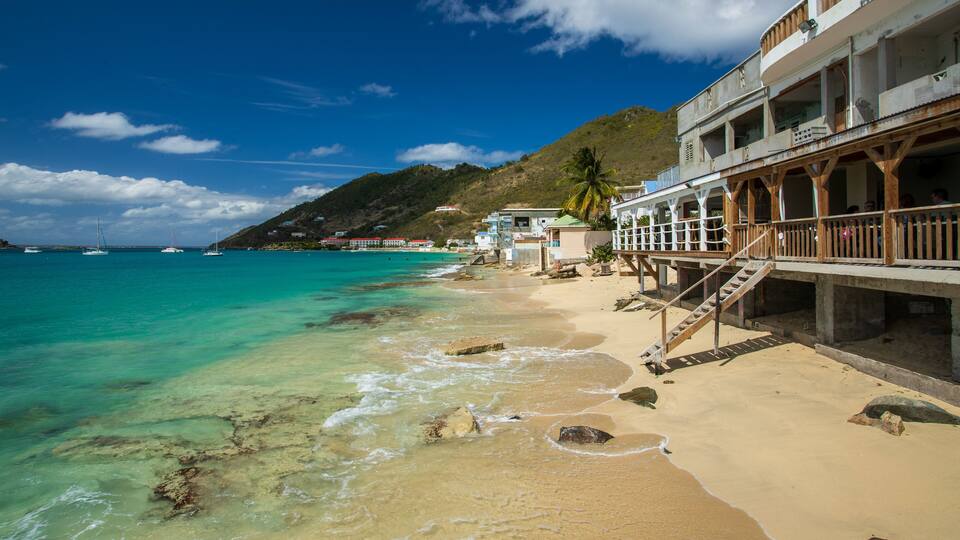 A beach with turquoise water, a wooden pier, and buildings on a hillside.