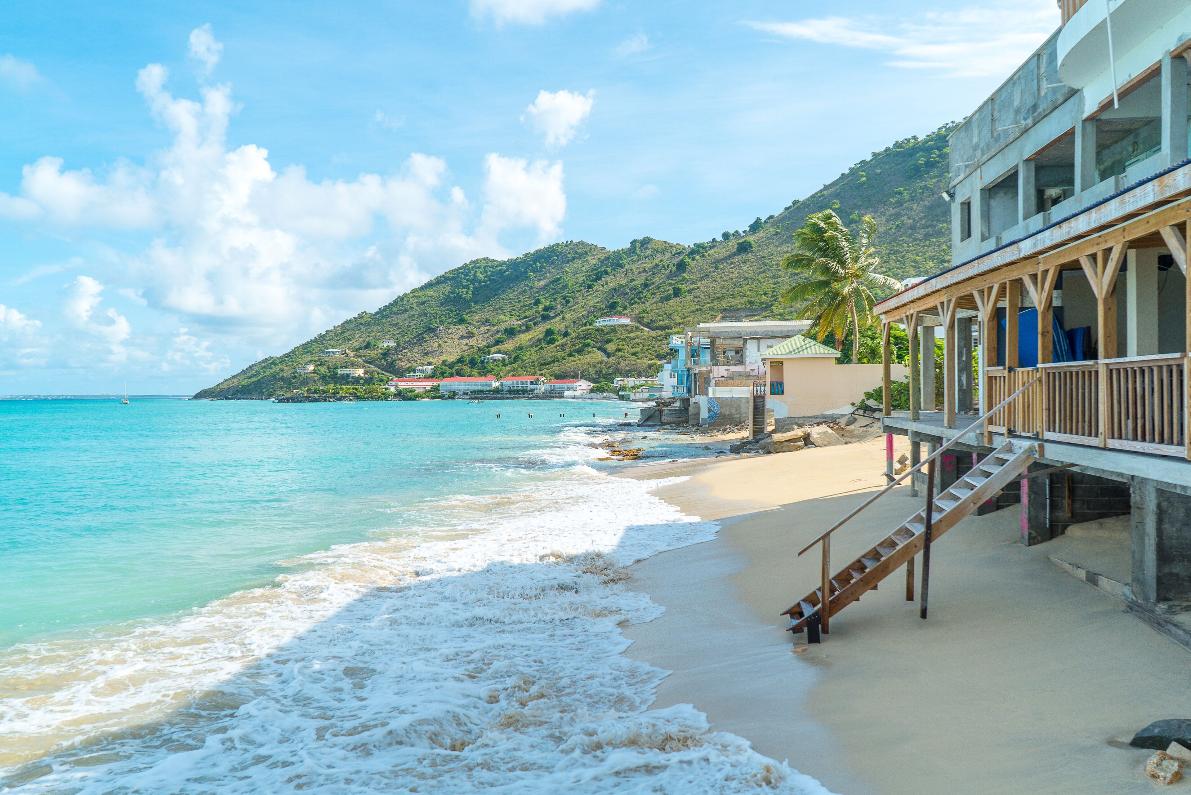 A beach with clear turquoise water, a wooden staircase leading to a building, and a hill with greenery in the background.