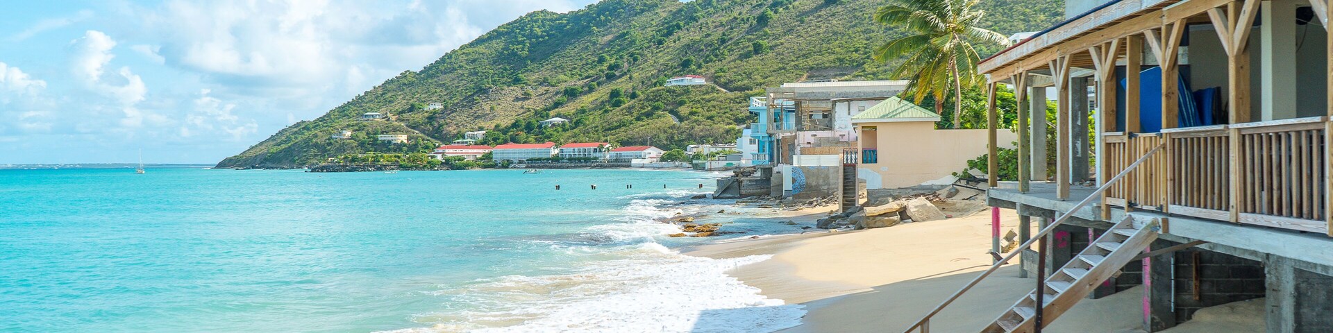 A beach with clear turquoise water, a wooden staircase leading to a building, and a hill with greenery in the background.