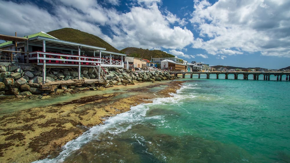 Une zone côtière caractérisée par un rivage rocheux, des eaux turquoise et limpides, une jetée s’avançant dans la mer et des bâtiments dotés d’un toit végétalisé, situés près du rivage.