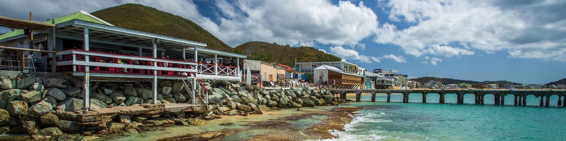 A coastal area with a rocky shoreline, clear turquoise waters, a pier extending into the sea, and buildings with a green roof near the shore.