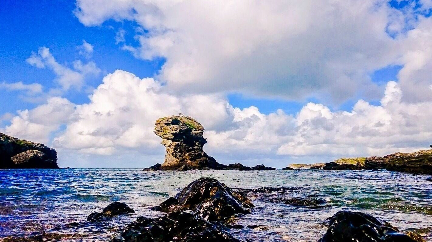 A coastal landscape with a prominent rock formation rising from the sea, surrounded by smaller rocks and cliffs.