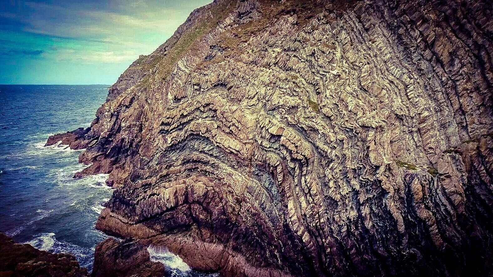 A rugged cliff with striations and a turbulent sea at its base.