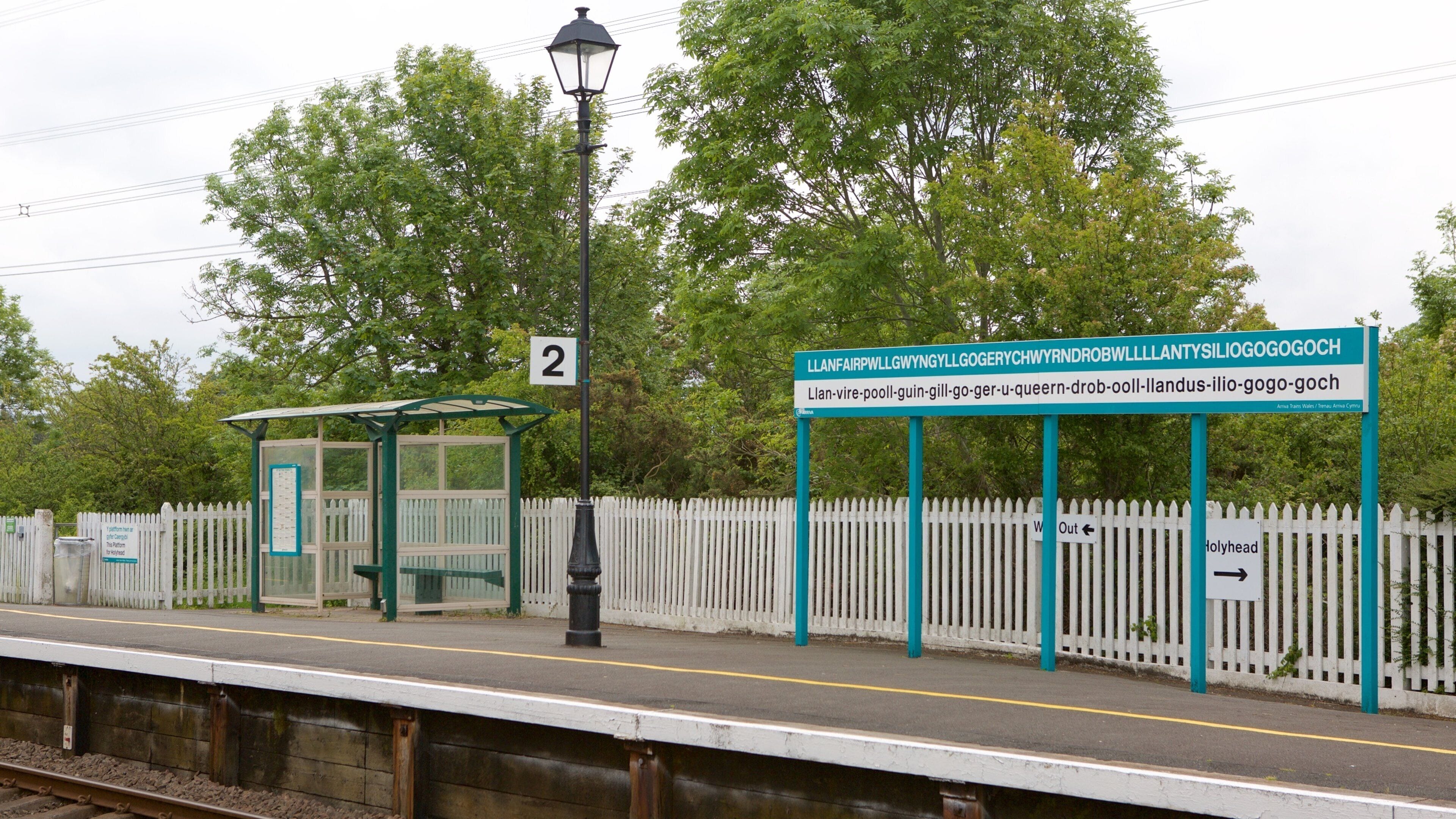 A train station platform with a shelter, a sign with Welsh text, and a white picket fence.
