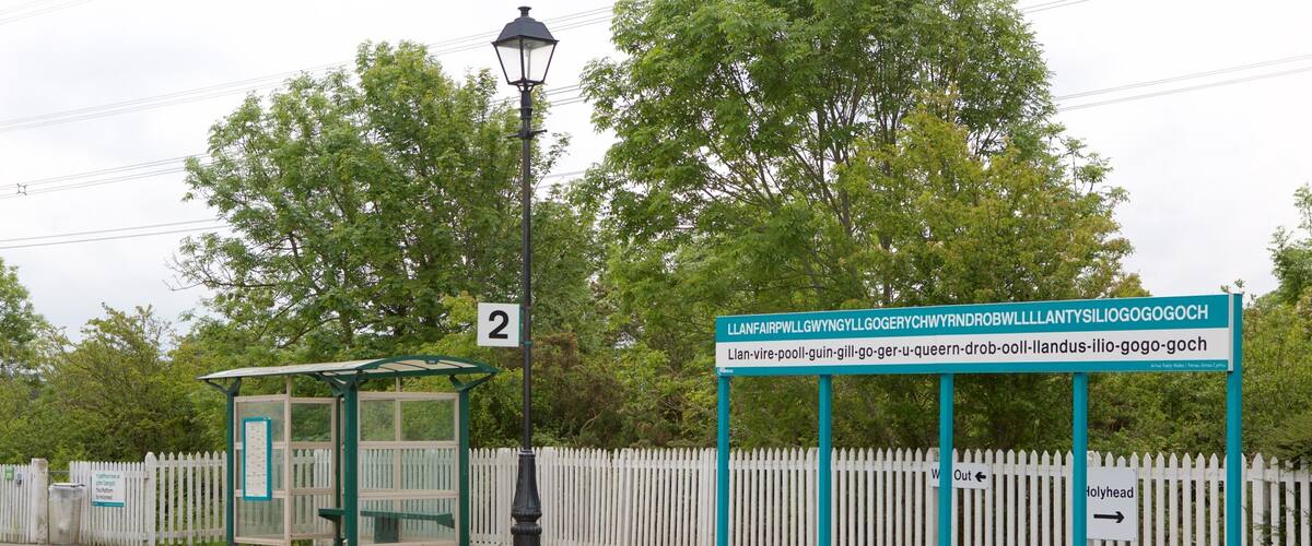 A train station platform with a shelter, a sign with Welsh text, and a white picket fence.