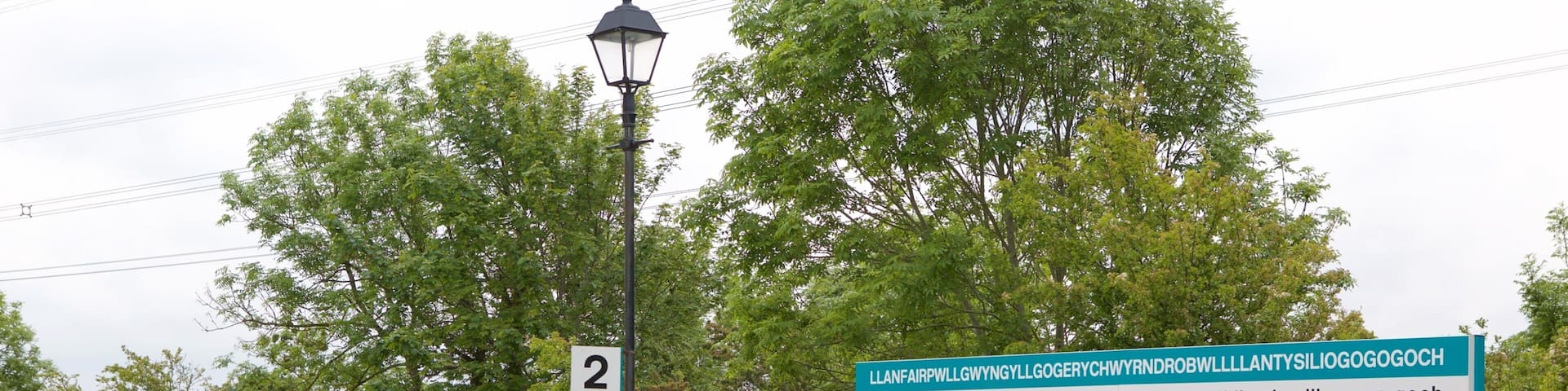 A train station platform with a shelter, a sign with Welsh text, and a white picket fence.