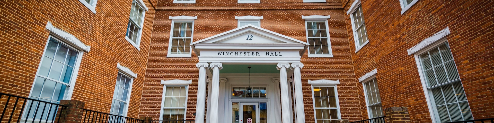 Un edificio de ladrillo con una entrada central, columnas y el nombre 'Winchester Hall' sobre la puerta.