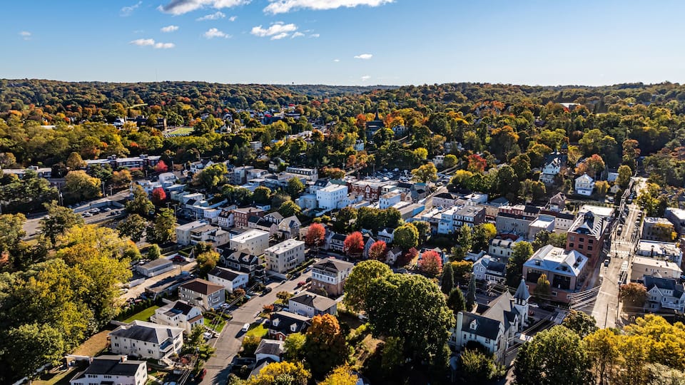 An aerial view of a residential area with houses, streets, and a hilly landscape in the background.