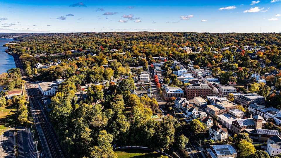 An aerial view of a town with dense tree coverage, a waterfront, and a mix of residential and commercial buildings.