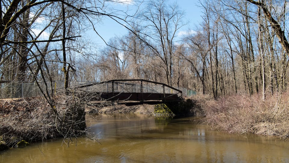 A rustic metal bridge over a murky river, surrounded by leafless trees and a fence.