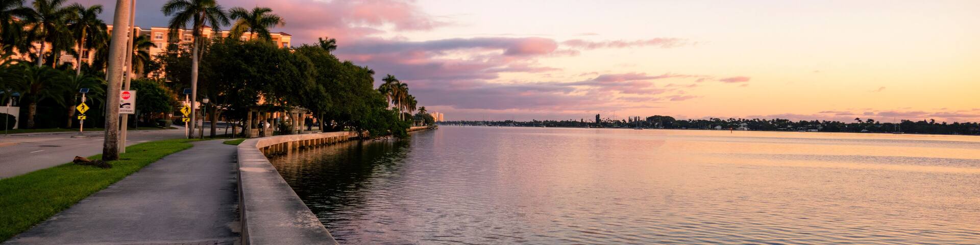 A waterfront promenade with palm trees, a stop sign, and a calm river reflecting the sunset sky.