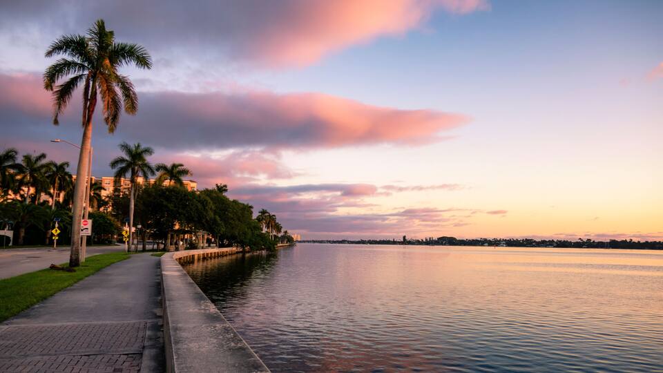 A waterfront promenade with palm trees, a stop sign, and a calm river reflecting the sunset sky.