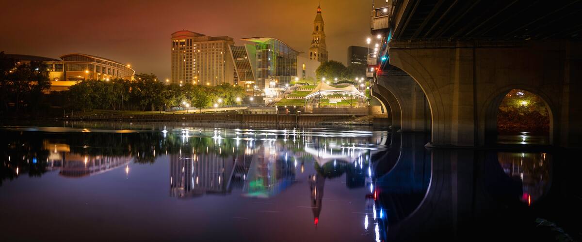 A nighttime cityscape with a river reflecting lights, a bridge, and a prominent tower.