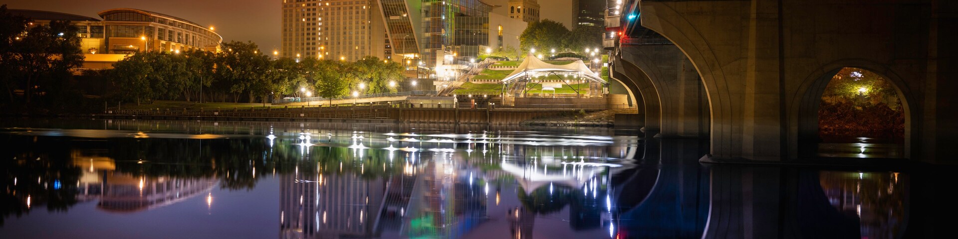 A nighttime cityscape with a river reflecting lights, a bridge, and a prominent tower.