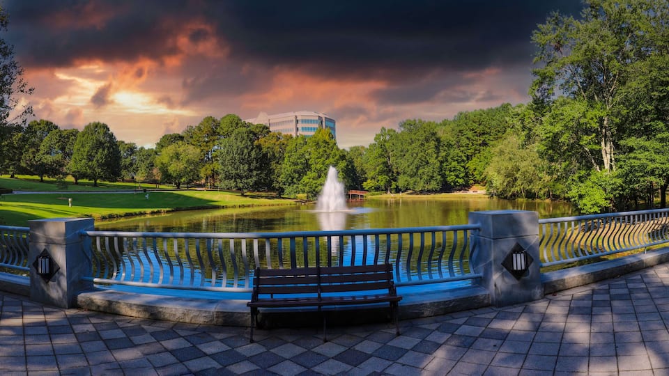 A park with a fountain, trees, a golf course, and a building in the background.