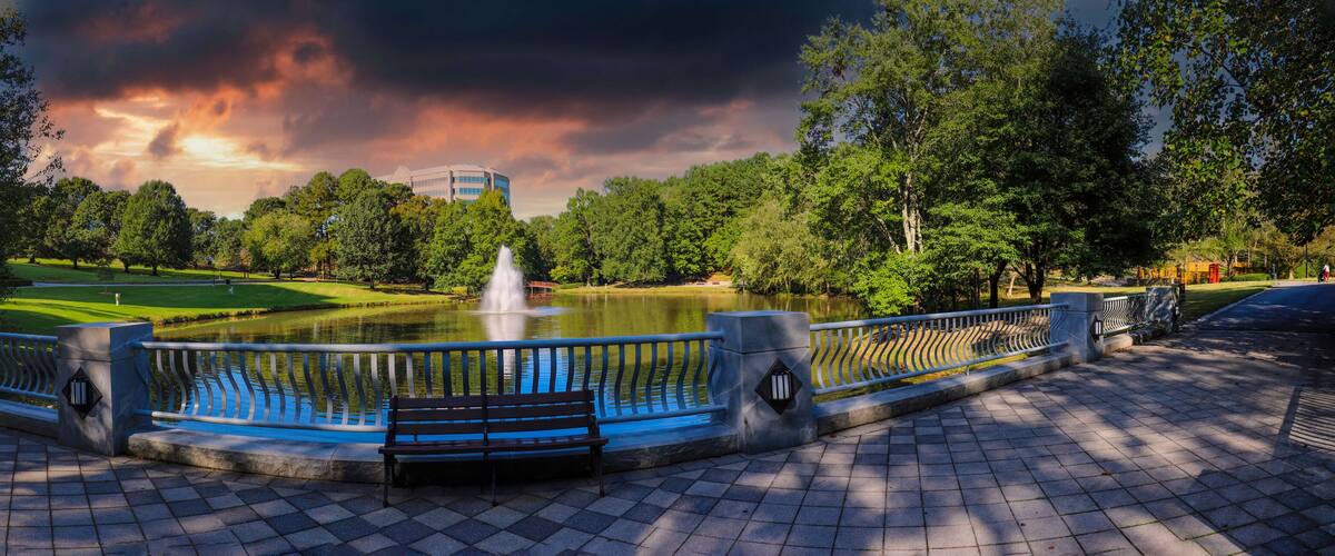 A park with a fountain, trees, a golf course, and a building in the background.