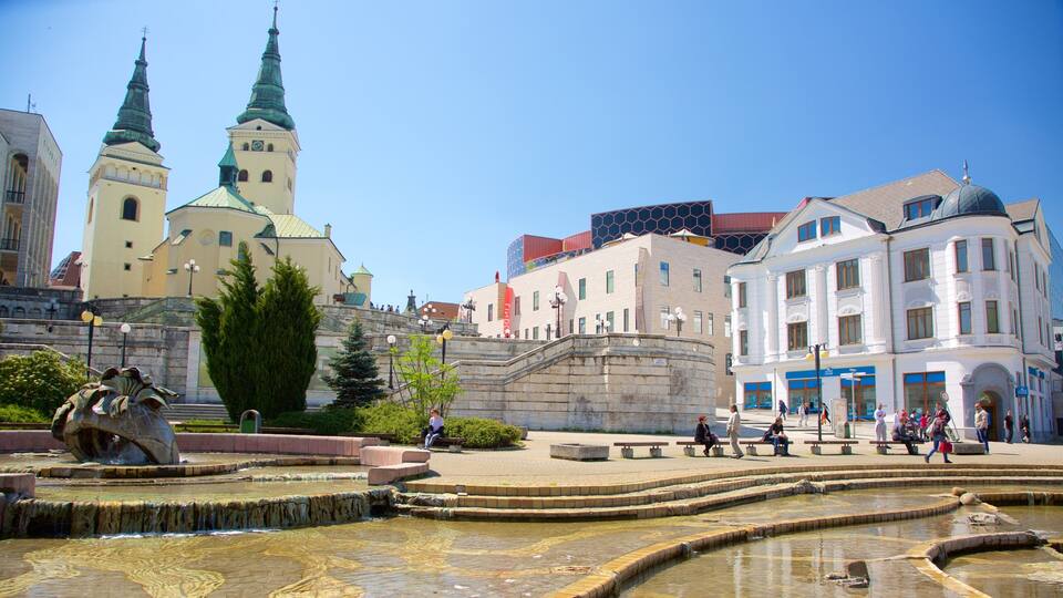 A historic square with a fountain, a statue, and a church with two spires.