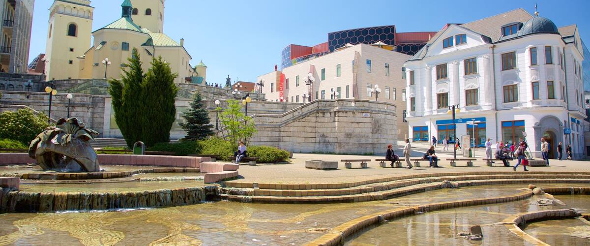 A historic square with a fountain, a statue, and a church with two spires.