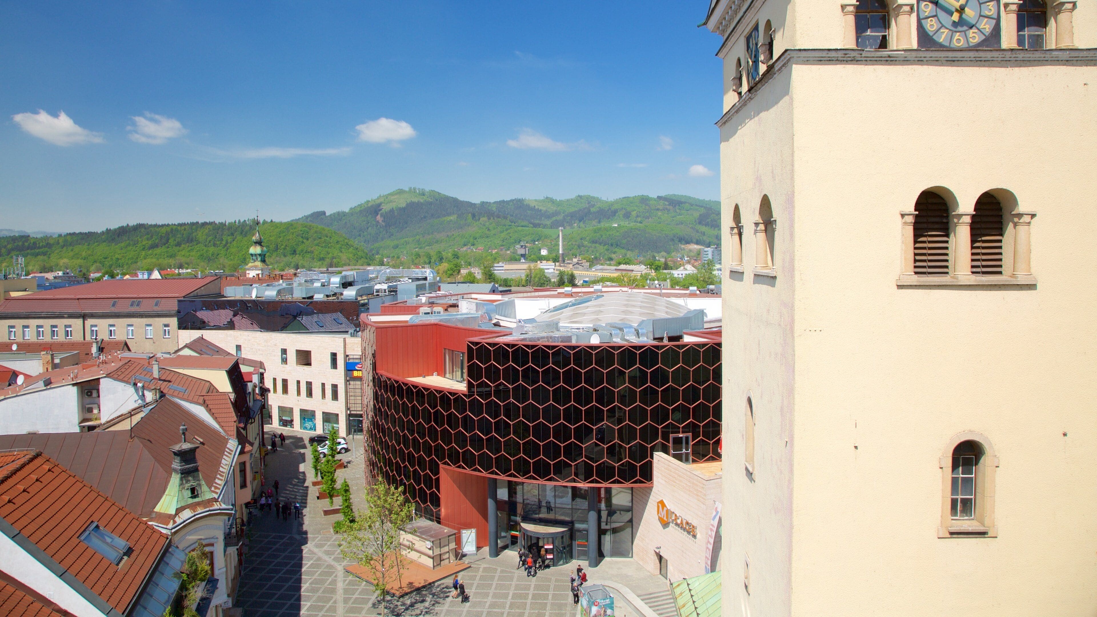 A cityscape with a prominent clock tower, a modern building with a hexagonal facade, and a mountainous backdrop.