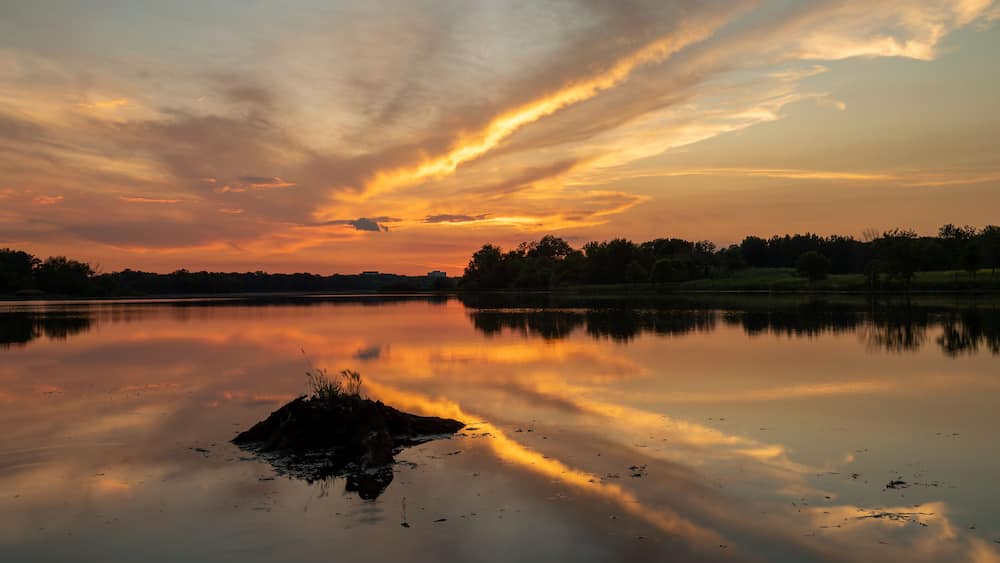 A sunset over a calm lake with a silhouette of trees and a rocky outcrop in the foreground.